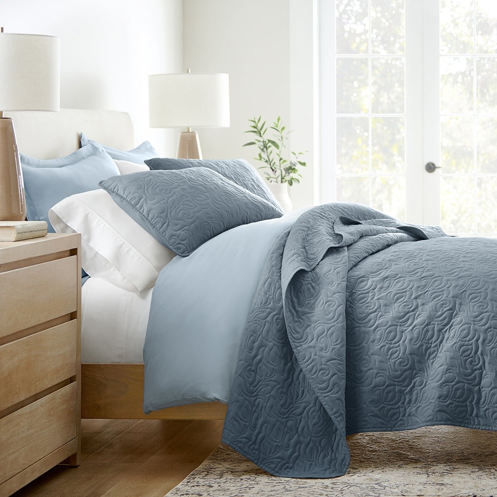 A bedroom features a bed with light blue sheets, textured blue-gray quilt and pillows, flanked by two wooden nightstands with lamps. A plant adds a touch of greenery near the windows.