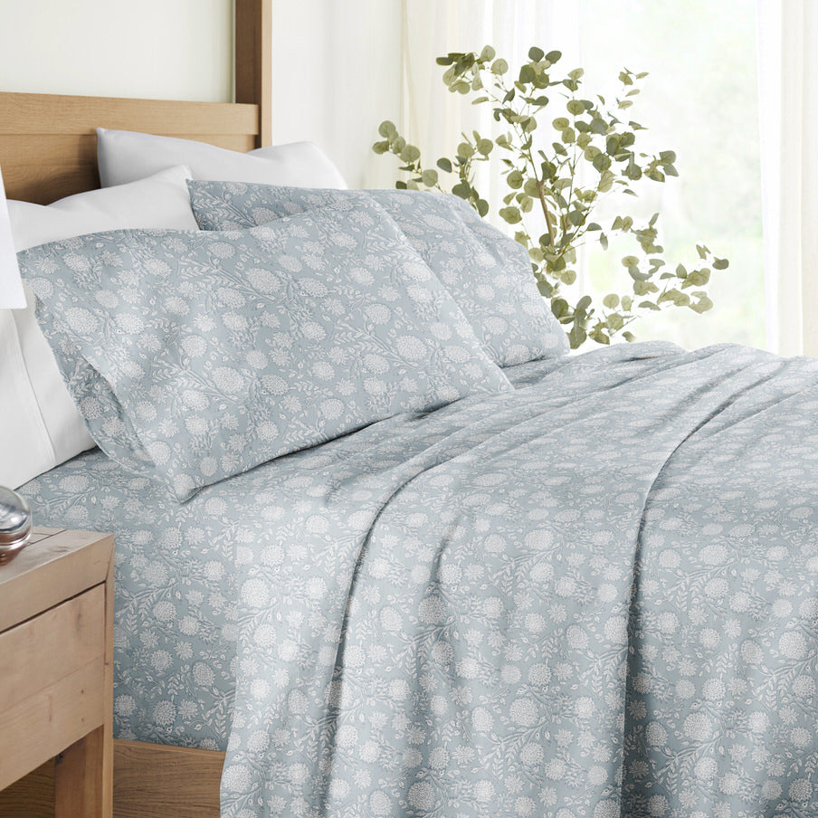A bed made with light blue floral-patterned sheets and pillowcases, set against a wooden headboard. There's a leafy plant next to the bed, enhancing the serene setup.
