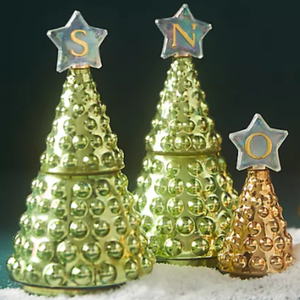 Gold and green glass jar candles shaped like Christmas trees, adorned with star toppers featuring letters \“S,\“ \“N,\“ and \“O.\“
