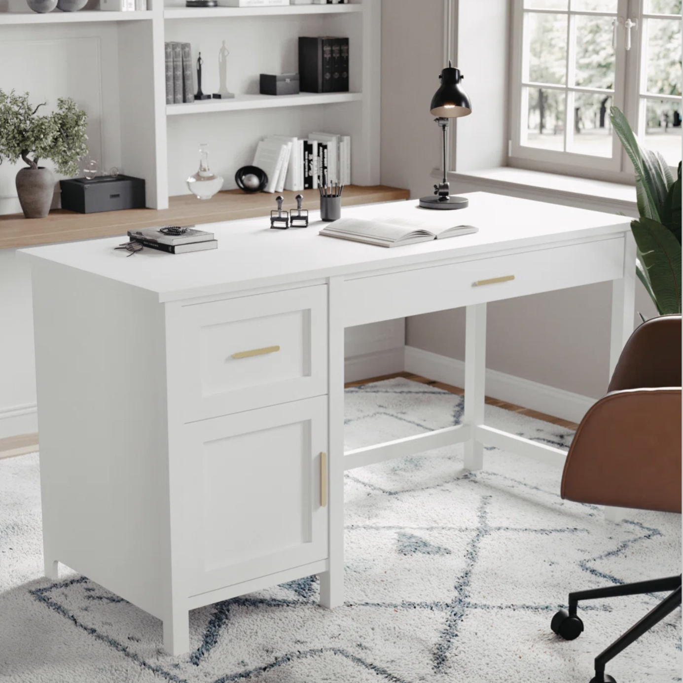 A white desk with gold handles features a drawer and cabinet, set in a home office with a lamp, a brown chair, and decorative items on shelves. The decor includes books, plants, and a modern rug on a wooden floor.