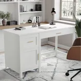 A white desk with gold handles features a drawer and cabinet, set in a home office with a lamp, a brown chair, and decorative items on shelves. The decor includes books, plants, and a modern rug on a wooden floor.
