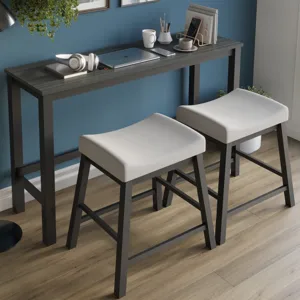 A sleek counter-height table set with two cushioned stools features a modern, minimalist design. The tabletop holds a laptop, headphones, and stationery, set against a blue accent wall with framed art.