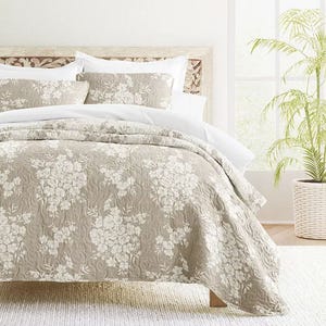 A bed is adorned with a floral-patterned beige quilt and matching pillow shams, surrounded by white pillows, all set against a wooden headboard with a carved design, and complemented by a potted plant by the window.