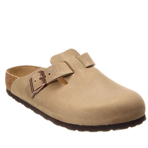 A tan suede slip-on clog with a closed toe and an adjustable buckle strap, featuring a cork footbed and dark brown sole.