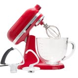 Red stand mixer with a glass bowl, featuring a whisk attachment installed. Also shown are a paddle and a dough hook attachment.