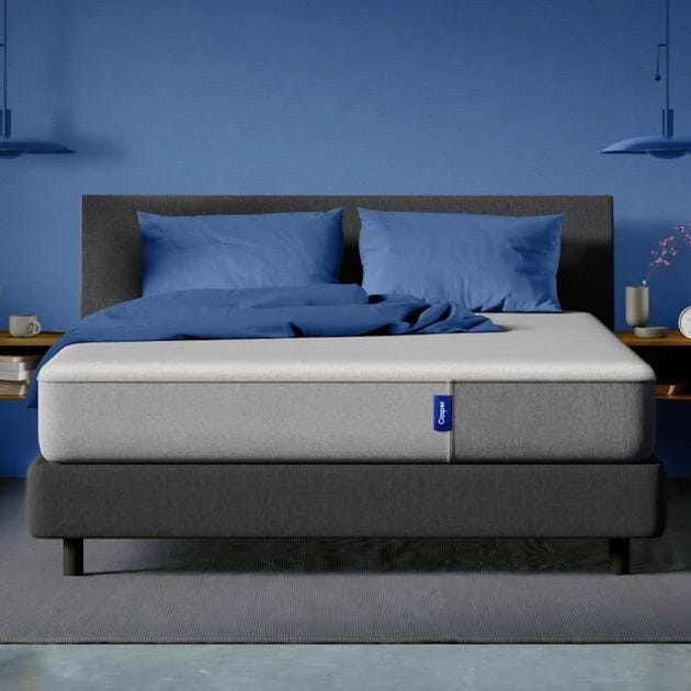 A gray upholstered bed with a thick, light gray mattress, blue bedding, and two blue pillows. The room features blue walls, hanging lights, and small bedside tables with decor.