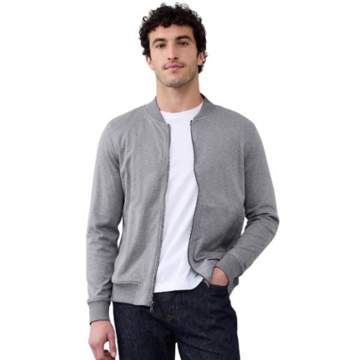 A man is wearing a casual outfit with a gray zip-up jacket over a white T-shirt and dark jeans. The style is simple and relaxed.