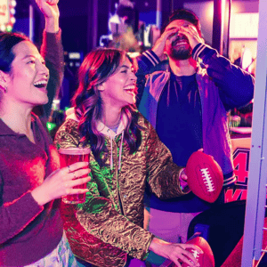 Three people are having fun at an arcade, with one holding a drink and the others engaging with a football-themed game, surrounded by colorful neon lights.