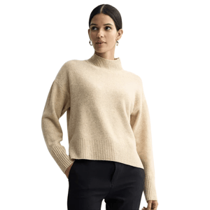A woman is wearing a beige high-neck sweater paired with black pants. She accessorizes with hoop earrings.