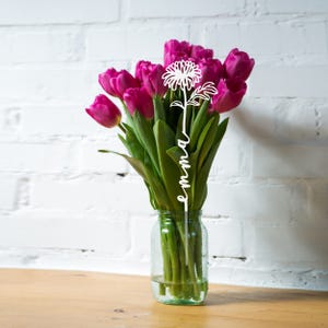 A bouquet of purple tulips in a glass vase features a custom metal flower decoration with a stylized daisy and the name \“emma\“ written vertically on the stem.