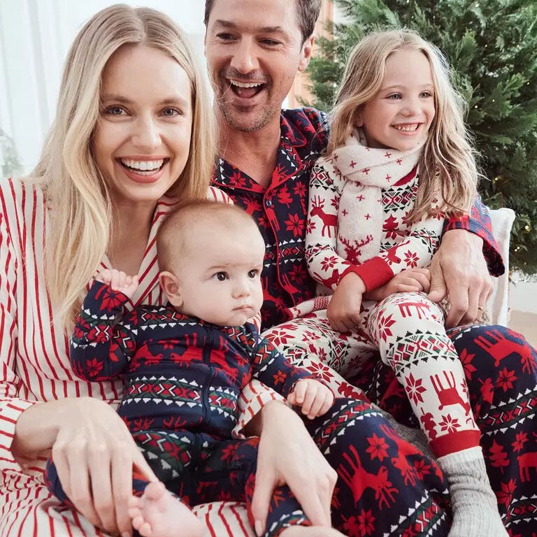 A family of four is wearing festive Hanna Andersson holiday pajamas with red, white, and blue designs featuring reindeer and snowflakes. The pajamas come in different patterns but coordinate in a holiday theme, perfect for family matching.