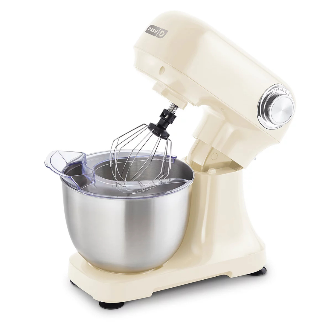 A cream-colored stand mixer with a stainless steel bowl, wire whisk attachment, and a transparent splash guard.