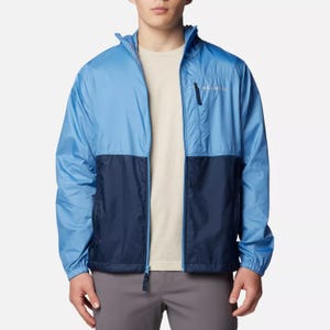 A man wearing a two-tone, blue and navy, lightweight windbreaker jacket with a zipper front and a logo on the chest.