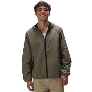 A smiling person is wearing a lightweight, olive green hooded jacket with black zipper details and beige pants.