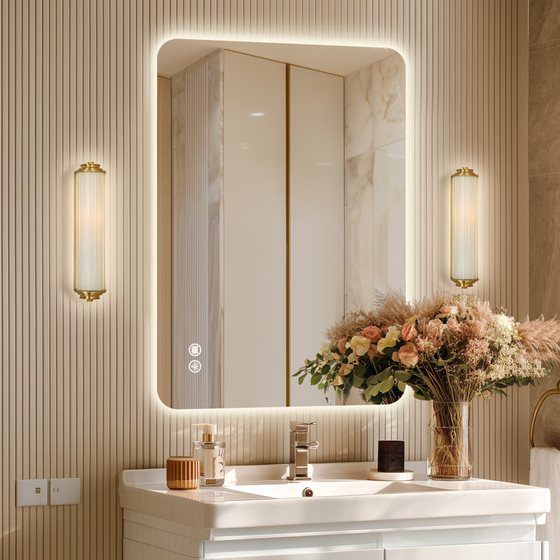 A bathroom scene featuring a backlit rectangular mirror with touch controls, modern wall sconces, a white sink, and a vase of flowers.