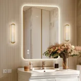 A bathroom scene featuring a backlit rectangular mirror with touch controls, modern wall sconces, a white sink, and a vase of flowers.