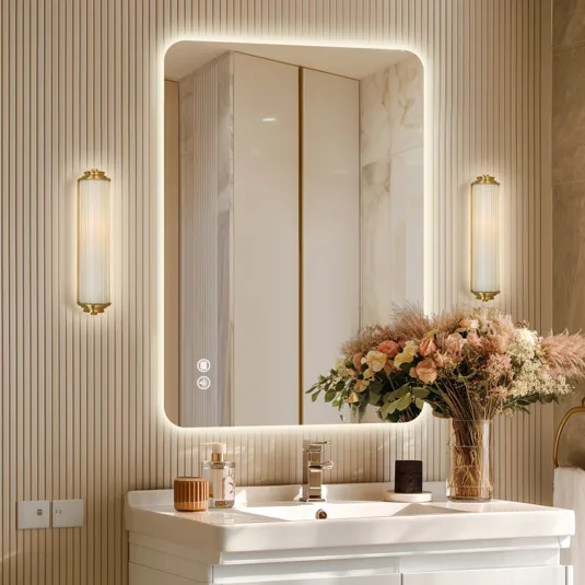 A bathroom scene featuring a backlit rectangular mirror with touch controls, modern wall sconces, a white sink, and a vase of flowers.