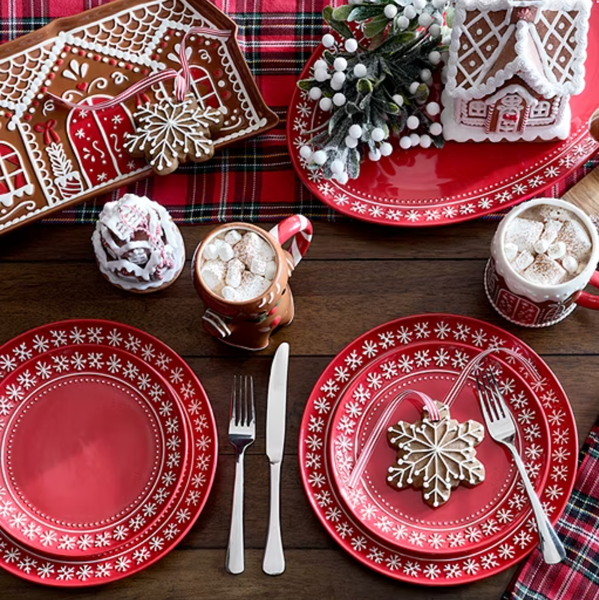 Festive red plates with white snowflake designs, gingerbread-themed mugs with hot cocoa, cookies shaped like snowflakes, and decorative gingerbread houses on a plaid tablecloth.