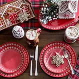 Festive red plates with white snowflake designs, gingerbread-themed mugs with hot cocoa, cookies shaped like snowflakes, and decorative gingerbread houses on a plaid tablecloth.