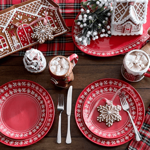 Festive red plates with white snowflake designs, gingerbread-themed mugs with hot cocoa, cookies shaped like snowflakes, and decorative gingerbread houses on a plaid tablecloth.
