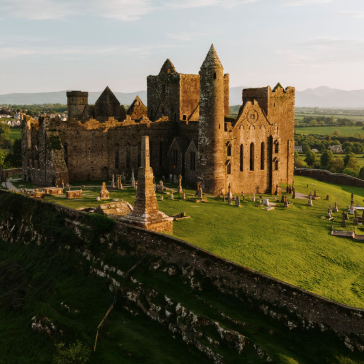 Historic stone castle ruins with cylindrical towers and arched windows, surrounded by old gravestones on a grassy hill overlooking a vast green landscape.
