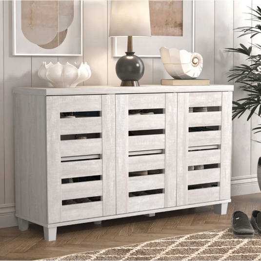 A wooden sideboard with slatted doors, featuring a light finish. It holds decorative items, including a lamp, a shell ornament, and a vase.
