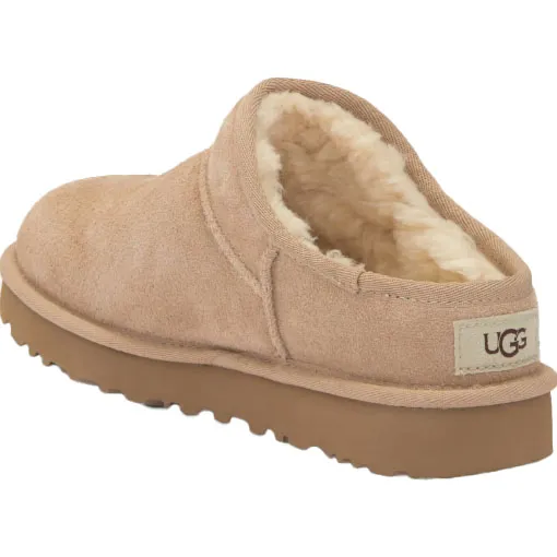 A beige UGG slip-on shoe with shearling lining and a brown rubber sole, featuring the UGG logo on the heel.