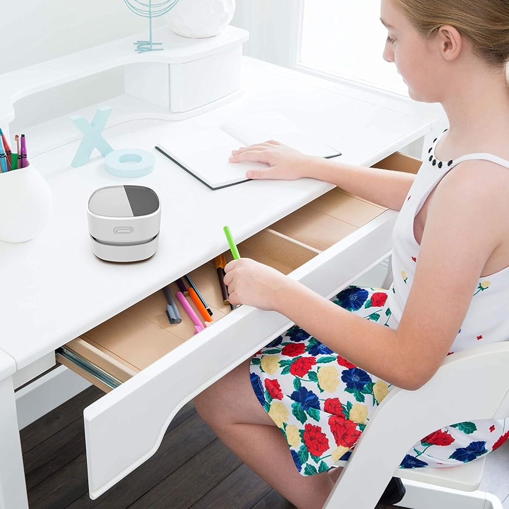 A young girl is drawing on a tablet at a white desk with a smart speaker and stationery holder nearby.
