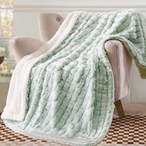 A plush, mint green and cream sherpa fleece blanket is draped over a beige armchair, set on a checkered brown and white rug.