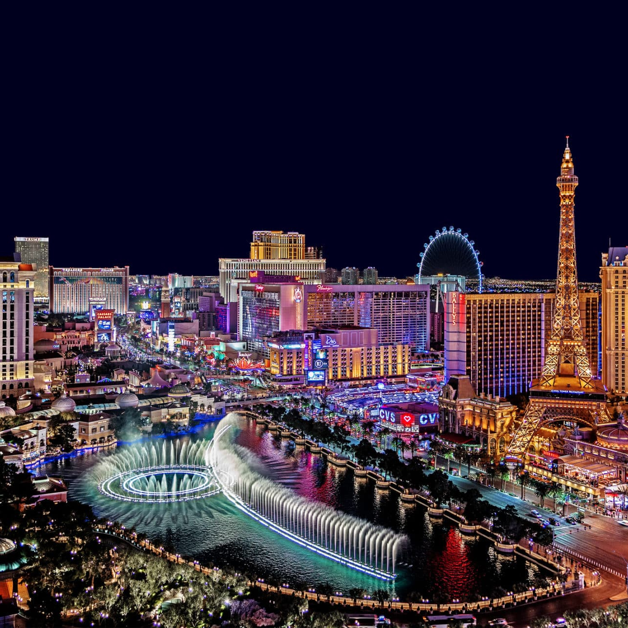 A nighttime view of the Las Vegas Strip features the Bellagio fountains, a replica of the Eiffel Tower, and brightly lit hotels and casinos, including a visible Ferris wheel and CVS store.
