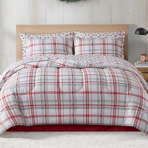 A cozy bed setup featuring a plaid comforter and matching pillow shams in red, green, and white hues. A small plant and book sit on a bedside table, with a holiday wreath on the wall.