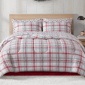 A cozy bed setup featuring a plaid comforter and matching pillow shams in red, green, and white hues. A small plant and book sit on a bedside table, with a holiday wreath on the wall.