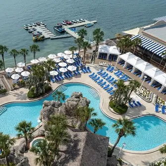 Aerial view of a coastal resort featuring a palm-lined pool area, with blue lounge chairs and umbrellas, alongside a marina with boats and watercraft.