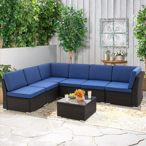 6-piece patio sectional sofa set with blue cushions on a dark wicker base, arranged in an L-shape around a square glass-top coffee table, set on a patterned outdoor rug in a garden setting with potted plants and decorative wall art.