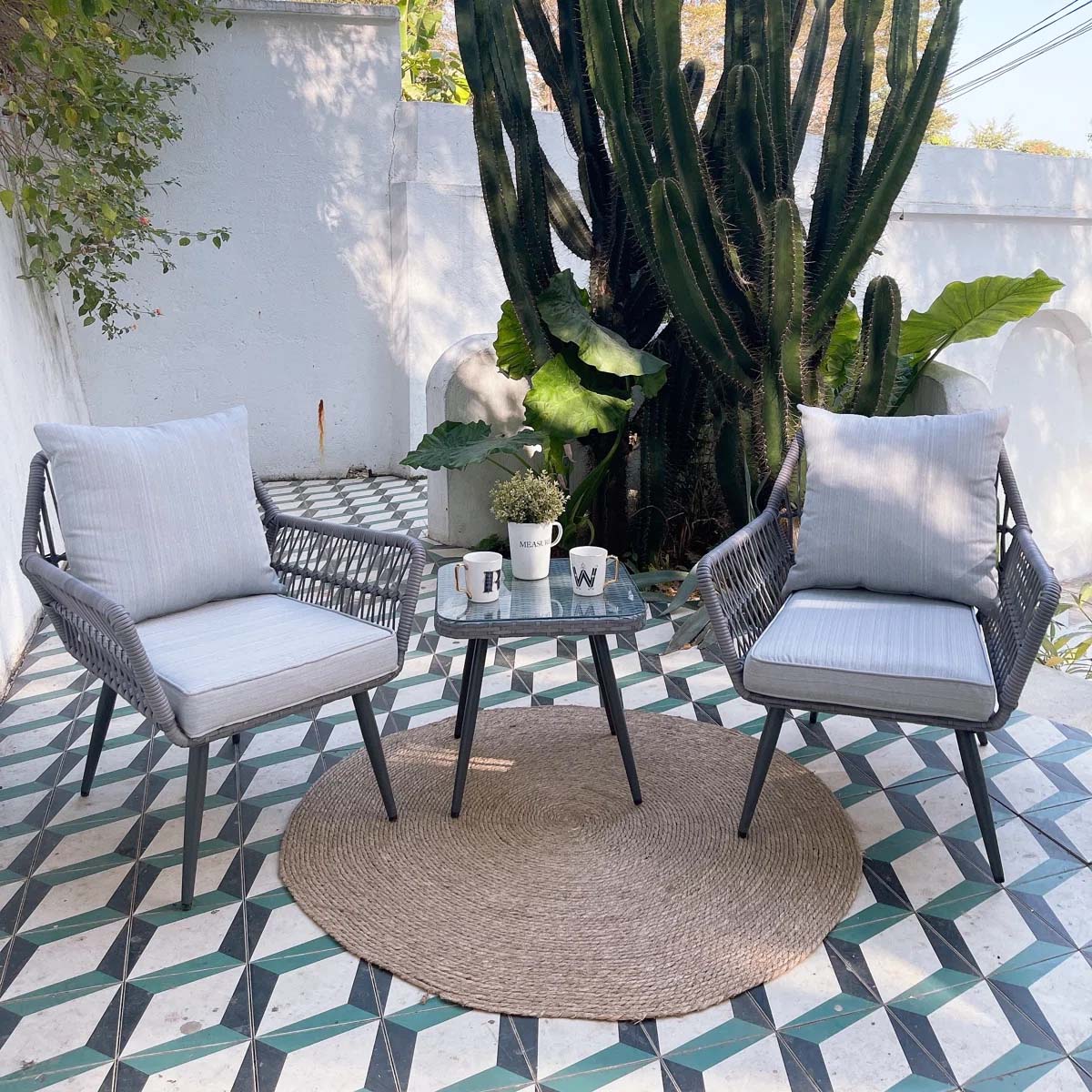 Outdoor patio scene with two gray cushioned chairs and a glass-topped table, set on a geometric-tiled floor with a jute rug, surrounded by lush plants and a large cactus.