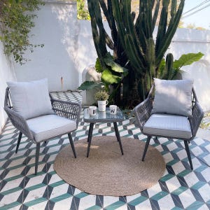 Outdoor patio scene with two gray cushioned chairs and a glass-topped table, set on a geometric-tiled floor with a jute rug, surrounded by lush plants and a large cactus.