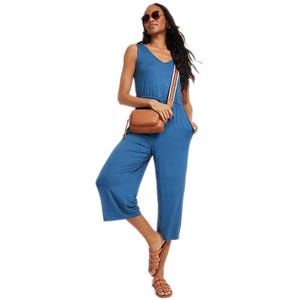 A woman wears a blue sleeveless V-neck, wide-leg jumpsuit paired with brown gladiator sandals, sunglasses, and a brown crossbody bag featuring a wide striped strap.