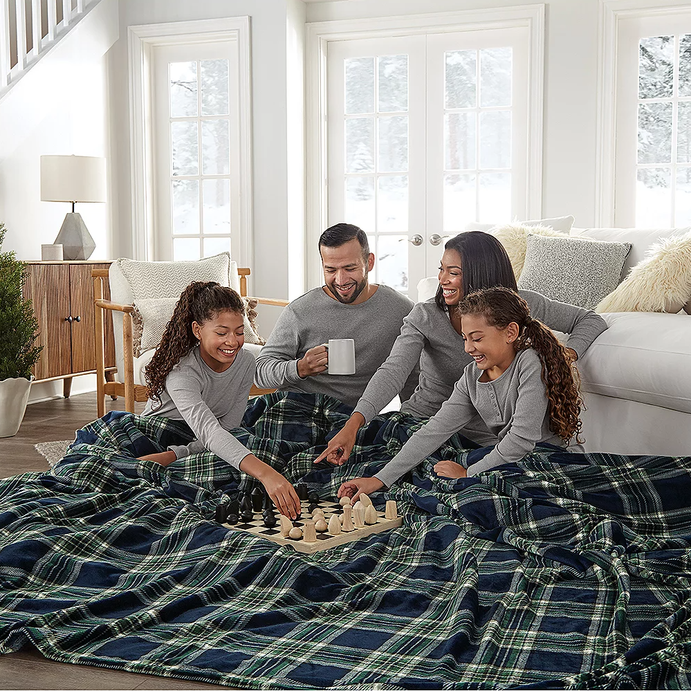 A large, plaid-patterned blanket measuring 120\“ x 98\“ is spread across a living room floor, where a family of four is gathered, playing chess. They are all dressed in matching gray loungewear, enjoying a cozy winter scene by the window.