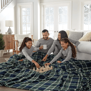 A large, plaid-patterned blanket measuring 120\“ x 98\“ is spread across a living room floor, where a family of four is gathered, playing chess. They are all dressed in matching gray loungewear, enjoying a cozy winter scene by the window.