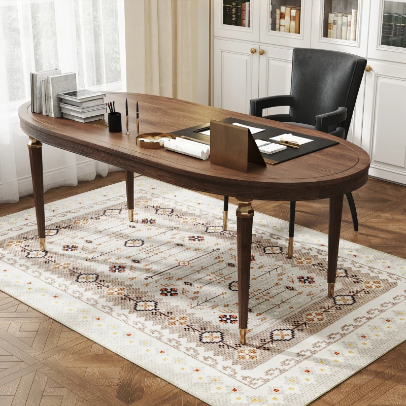 An oval wooden desk with brass accents sits on a patterned rug, accompanied by a black chair. The desk holds books, pens, and stationery items in an elegant office setting.