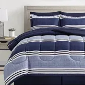 A bedding set with a striped pattern in navy and white is displayed on a bed with matching shams. The room includes a wooden headboard, side tables with lamps, and framed abstract artwork on the wall.