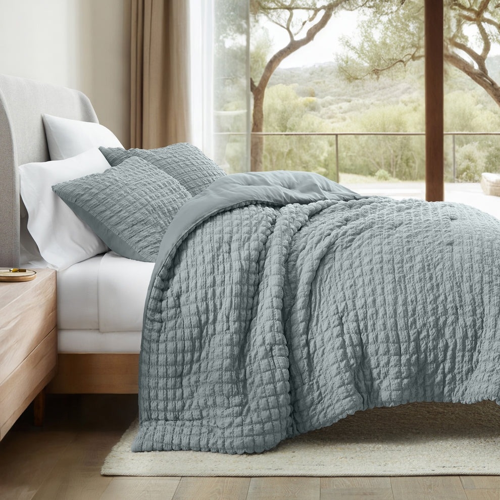 A bed with a textured gray quilt and matching pillowcases is shown, positioned next to a window overlooking a scenic outdoor view.
