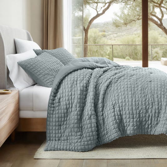 A bed with a textured gray quilt and matching pillowcases is shown, positioned next to a window overlooking a scenic outdoor view.