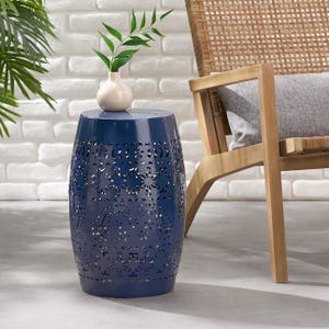 Blue metal garden stool with floral cutout design, topped with a small white vase holding green leaves, placed next to a wooden chair with a cushion, set against a white brick wall.
