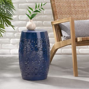 Blue metal garden stool with floral cutout design, topped with a small white vase holding green leaves, placed next to a wooden chair with a cushion, set against a white brick wall.