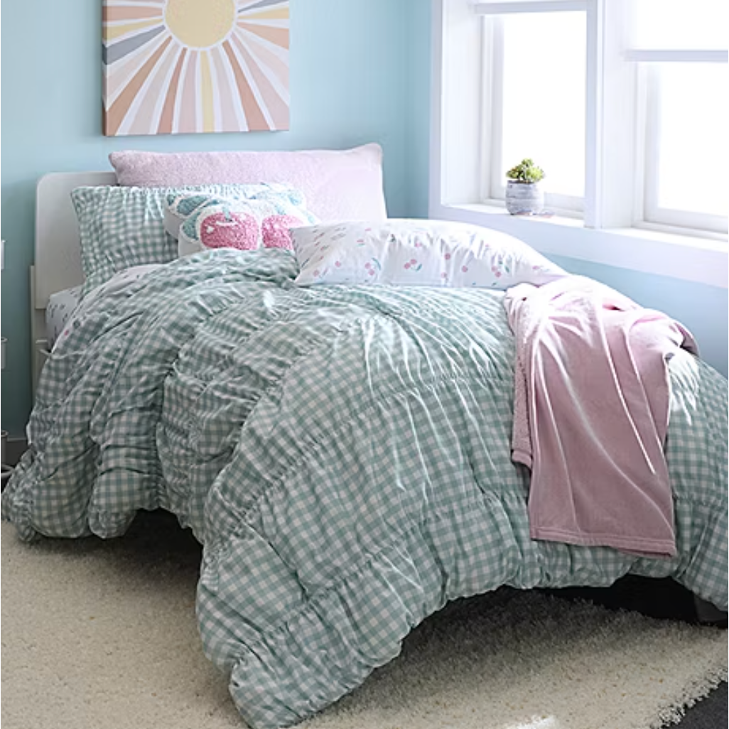 A cozy bedroom setting features a mint green gingham comforter, assorted decorative pillows including a pink llama design, and a pink blanket. A cheerful sun-themed canvas hangs above, and a small plant sits on the windowsill.
