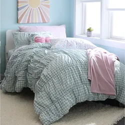 A cozy bedroom setting features a mint green gingham comforter, assorted decorative pillows including a pink llama design, and a pink blanket. A cheerful sun-themed canvas hangs above, and a small plant sits on the windowsill.