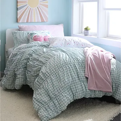 A cozy bedroom setting features a mint green gingham comforter, assorted decorative pillows including a pink llama design, and a pink blanket. A cheerful sun-themed canvas hangs above, and a small plant sits on the windowsill.