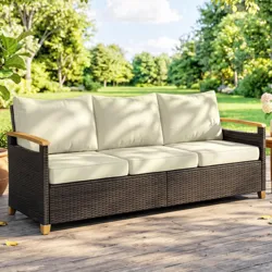 Outdoor wicker sofa with cream cushions and wooden armrests, set on a wooden deck in a garden setting.