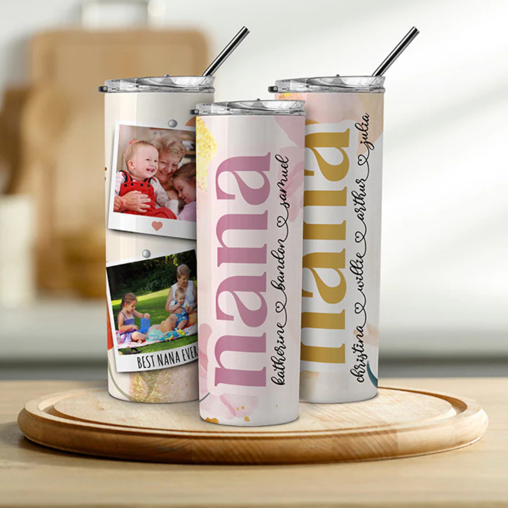 Three personalized tumblers with metal straws feature the word \“nana\“ and names in cursive. One tumbler showcases photos of children with a message, \“Best Nana Ever.\“ They're placed on a round wooden tray.
